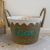 Een geweven mand met touwgrepen en een decoratieve witte bies staat op een vloer met sterrenpatroon. De naam Lucas staat in groenblauw schrift op de voorkant. Een deel van een wieg is zichtbaar op de achtergrond.