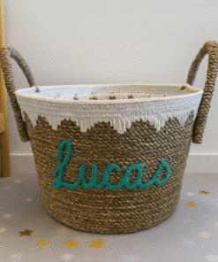 Een geweven mand met touwgrepen en een decoratieve witte bies staat op een vloer met sterrenpatroon. De naam Lucas staat in groenblauw schrift op de voorkant. Een deel van een wieg is zichtbaar op de achtergrond.