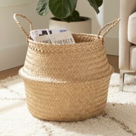 De COCO - lichte opbergmand van zeegras met twee handgrepen zit op een crèmekleurig tapijt en bergt tijdschriften op. Een plant en een deel van een beige sofa verschijnen op de achtergrond.