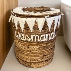 De INDRA decoratieve wasmand van donker zeegras, met de tekst 'wasmand' en wit zigzagpatroon, staat op de badkamervloer naast het bad en een houten wandpaneel.