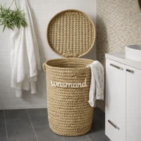 De RIVA - wasmand van palmblad met deksel staat open in een badkamer naast witte handdoeken, een plant en een witte kast, met kleding gedrapeerd over de rand.