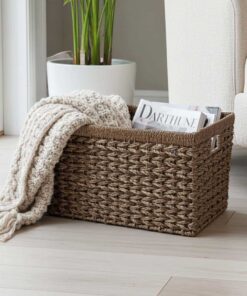 De Elva decoratieve opbergmand van zeegras staat op een lichthouten vloer naast een crèmekleurige bank. Binnen liggen een opgevouwen gebreide crèmekleurige deken en tijdschriften, met een potplant zichtbaar op de achtergrond bij het raam.