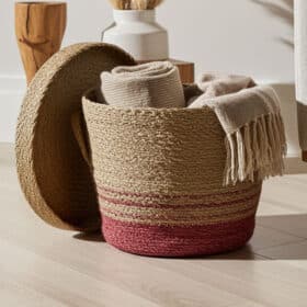 De RODO - ronde zeegras mand met deksel, met een roodroze basis, staat open op een lichte houten vloer met een beige handdoek en een deken met franjes erin. Op de achtergrond staan nog een mand en een vaas.